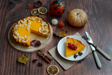 Bright orange pumpkin pie on a wooden stand and a white plate, cinnamon sticks, small pumpkins and autumn foliage
