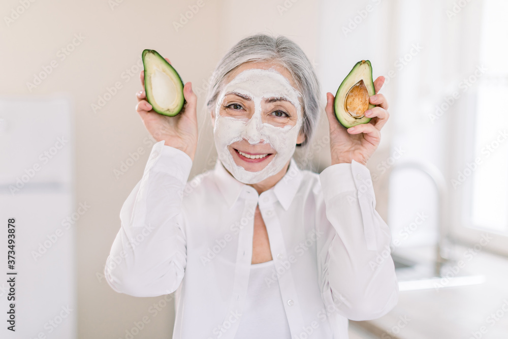 Wall mural portrait of smiling attractive elderly woman holding two halves of avocado, with nourishing facial c