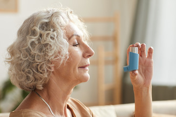 Side view close up portrait of senior woman using inhaler for asthma or breathing problems in home setting - obrazy, fototapety, plakaty