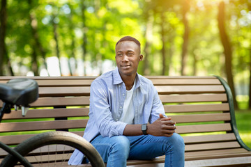 Handsome African American man with modern bicycle relaxing on bench at park