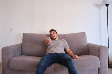 Portrait of a bored and tired young man sitting on a sofa watching television and changing channels with the remote control on his hands.