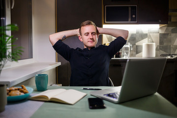 Tired businessman sitting at the table at home, modern cozy house, looking at the laptop screen, feels pleased with pride for the work done, calm person is resting, hands resting behind his head
