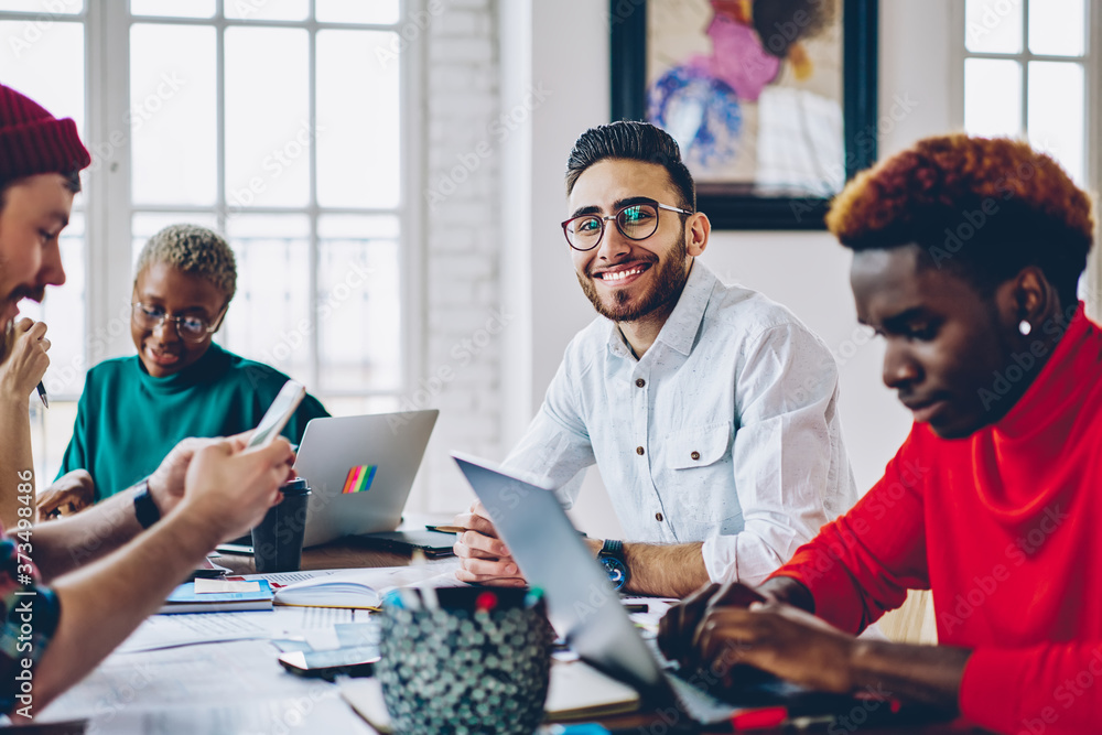 Poster portrait of happy male software developer sitting at meeting table with diverse colleagues and smili