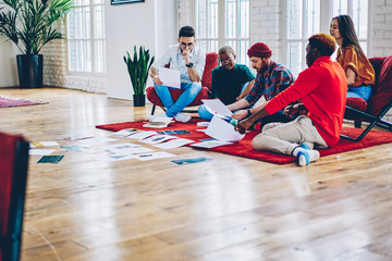 Diverse group of male and female designers cooperating on paper sketches sitting in trendy apartment, multicultural hipster people planning strategy of collaborative process in modern loft interior