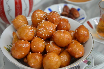 Traditional oriental dessert speciality Legematt (Luqaimat), fried dough balls in a porcelain bowl, blurred dates and a cup of tea behind, blurred background
