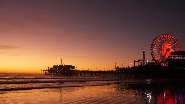 Twilight waves against classic illuminated ferris wheel, amusement park on pier in Santa Monica pacific ocean beach resort. Summertime iconic symbol of California glowing in dusk, Los Angeles, CA USA