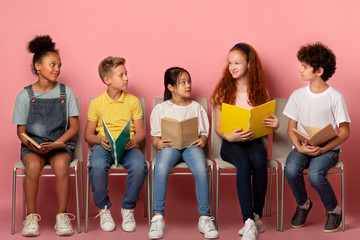 Classmates of different races with school supplies looking at each other on chairs over pink...