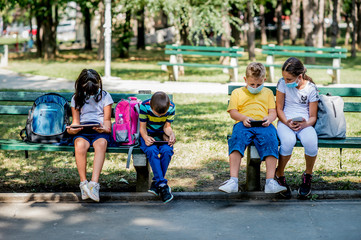 Back to school during quarantine. Children with backpacks wearing protective face mask and using mobile phones and digital tablets.