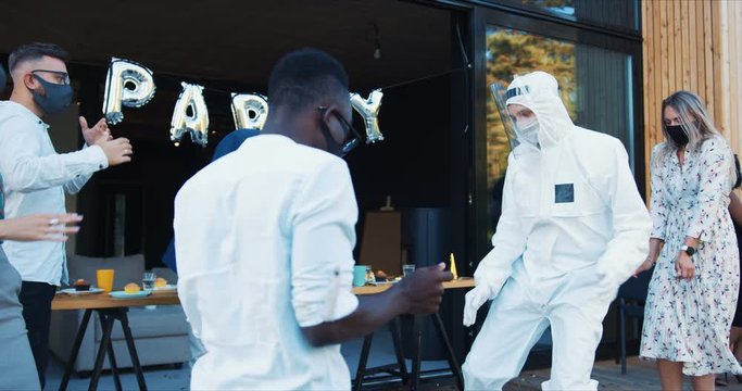 Return to normal life after COVID-19. Young happy friends dance in safety costumes and masks at fun party slow motion.