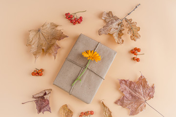 Autumn flat lay composition. Pretty gift box wrapped with craft paper and decorated with flower on orange background among dry foliage. Thanksgiving day concept. Copy space. Selective focus