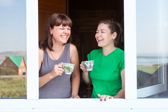 Two adult girlfiends laughing in countryside house window frame, women drinking morning coffee together