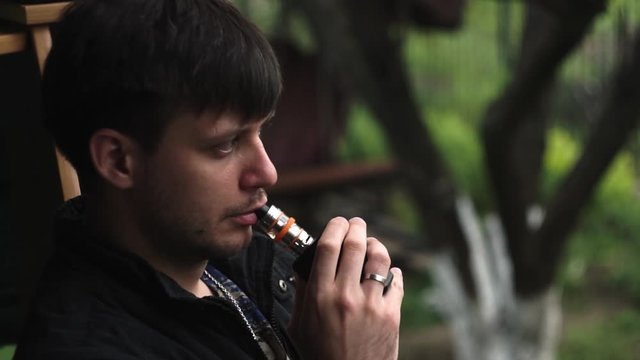 Vape man. Young white caucasian guy in a black jacket smoking and releasing steam from an electronic cigarette in the backyard in the summer. Bad habit. Vape activity.