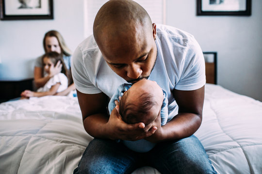 Father kissing newborn on bed with mother and daughter in background