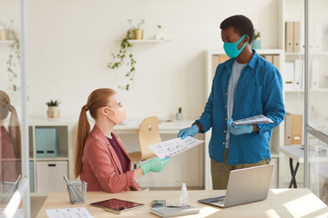 Portrait of young businesswoman wearing mask and gloves handing documents to African-American colleague while working in post pandemic office, copy space