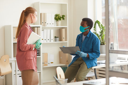 Portrait of young businesswoman wearing mask and gloves talking to African-American colleague while working in post pandemic office