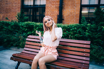 Cheerful hipster girl in electronic earphones listening received audio message with positive...