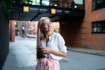 Obraz premium Pretty hipster girl in classic glasses for eyes protection standing at urban setting pondering on blog idea, attractive pink haired millennial woman with cellphone technology thinking outdoors