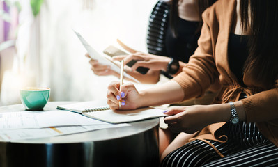 Close-up of hand young asian businesswoman holding pencil writing on book while she working with friend and blurred her friend holding smart phone and paperwork