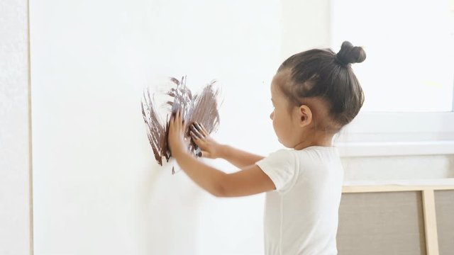 interested girl kid in white t-shirt draws on white canvas with brown paint on hands standing at atelier wall under bright sunlight