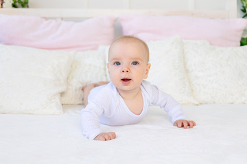 baby girl 6 months old in a white bodysuit lying on her stomach on a white bed at home