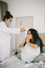 Female sneezing and covering her face with a tissue while having her fever checked by a nursewith digital thermometer.