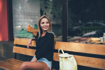 Caucasian cheerful hipster girl with perfect blonde hair taking rest on bench in city enjoying solo...