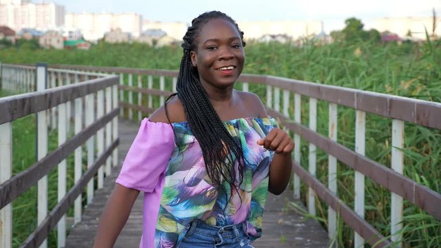 attractive young African-American woman with cornrows in bright color blouse dances on empty wooden bridge in green reeds against city slow motion