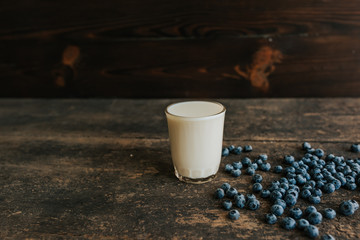 A transparent glass with milk. Blue fresh blueberries are scattered on an old brown wooden cracked table.