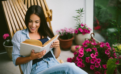 Young beautiful happy woman relaxing on cozy balcony, reading a book,