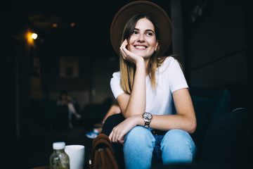 Fototapeta na wymiar Cheerful joyful caucasian female hipster girl satisfied with free time in cafe interior resting, attractive 20s woman in stylish hat laughing and having fun indoors dressed in cool trendy apparel