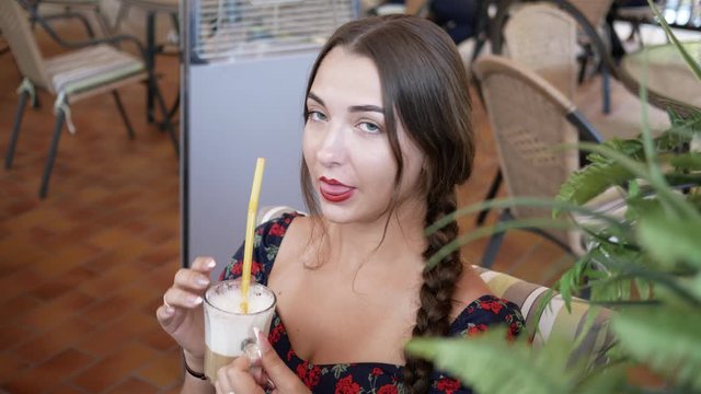 young beautiful woman with red lipstick on lips drinks coffee through straw in outdoor summer cafe on terrace