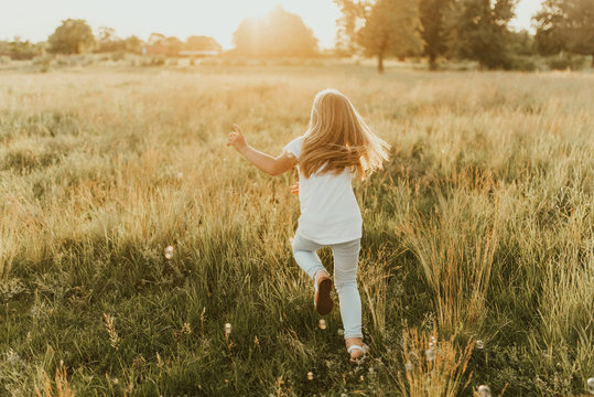 a little blonde girl dressed in light-colored clothes and a white T-shirt runs away from soap bubble. Along the tall green grass towards the setting sun. Warm summer sunny weather at sunset.