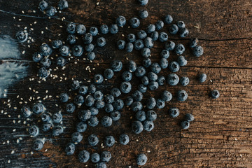 Blue fresh blueberries are scattered on an old brown wooden cracked table. Sesame seeds were poured between the fruits. Wet drops of milk on the table. basil leaves, green fresh seasoning.