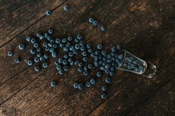 Fresh blue matte round blueberries filled in a transparent glass stands on a wooden brown shabby textured table and berries are scattered everywhere