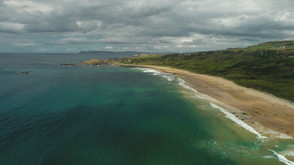Fototapeta premium Ireland sand beach aerial view: ocean waves, sandy coastline, white shore with greenery meadows. Epic Irish landscape with gray clouds on sky in summer day cinematic shot