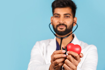 Indian / Asian doctor with a stethoscope and a red heart in his hands, in a white coat on a blue...