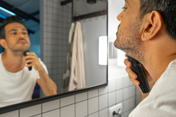 handsome man is shaving his beard with trimmer machine in front of bathroom mirror