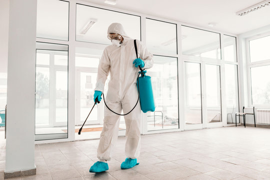 Worker in white sterile uniform, with rubber gloves and mask on holding sprayer with disinfectant and sterilizing school.