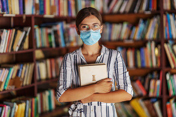Portrait of young attractive college girl standing in library with face mask on holding a book....
