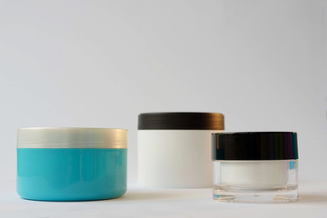 2 white jars with a black lid, one blue cream jar with a gray lid on a white background
