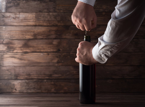 The hands of an unrecognizable waiter in a white shirt opens a bottle of red wine with a corkscrew on a dark wood background with copy space. Winemaking concept.