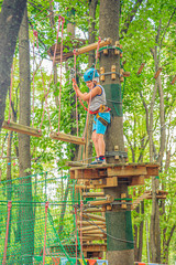 a teenage boy in equipment and a helmet stands on a round wooden platform and holds on to a rope in Outdoor Adventure forest park