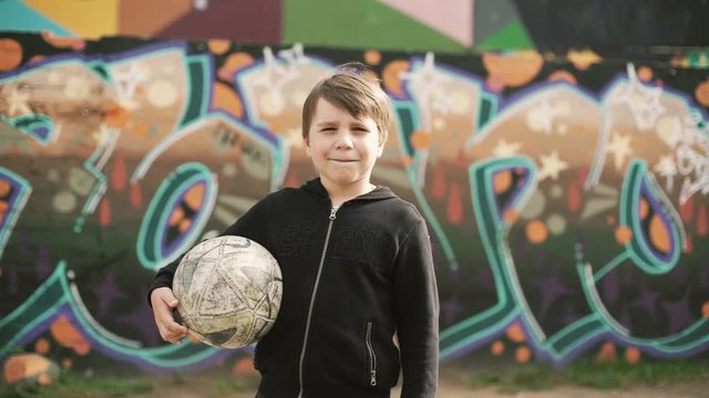 a little boy stands with a soccer ball in his hands against the background of a wall with graffiti, looks at the camera and makes funny faces