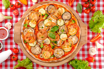 Vegetarian tasty pizza with vegetables, zucchini, eggplant, mushrooms, slices of tomato, fresh basil and tomatoes sauce on a wooden board, rustic village background, top view.
