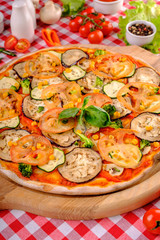 Vegetarian tasty pizza with vegetables, zucchini, eggplant, mushrooms, slices of tomato, fresh basil and tomatoes sauce on a wooden board, rustic village background, top view.