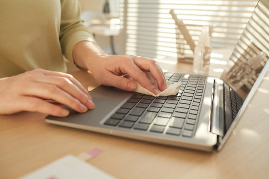 Close up pf unrecognizable woman sanitizing laptop while working at desk in post pandemic office lit by sunlight, copy space