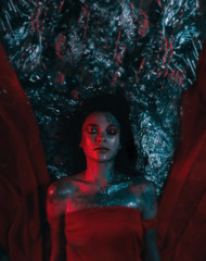 Fototapeta premium Glittering goddess in the red dress lies in the water, illuminated by blue and red light