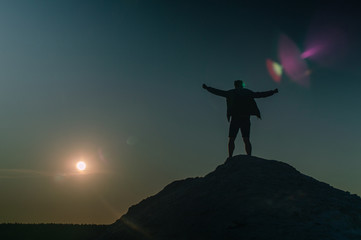 Silhouette of happy man on the mountain peak, spreads arms and looks at the sunset