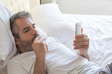 One man looks thoughtfully into a mobile phone. The man lies on the bed and uses a mobile phone.