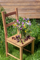 Bouquet of wildflowers and ripe cherries on a wooden chair in the grass, summer farm rustic concept. Natural background